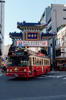 A vibrant red bus is driving through a city street, passing under a traditional Chinese-style gate with intricate blue and gold details. Surrounding the scene are modern urban buildings. The area seems to be a busy intersection, as there are traffic lights visible.