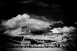 An expansive historical palace dominates the center beneath a dramatic sky filled with large, billowing clouds. The structure features layered architecture with multiple levels and terraces, constructed with a series of stepped walls. The foreground shows an open area with a few trees and people scattered, while the background consists of mountain ranges.