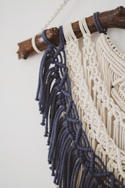 Detailed macramé wall hanging featuring dark blue and cream-colored cords intricately knotted. These are attached to a smooth wooden branch, creating a decorative piece with an artisanal feel.