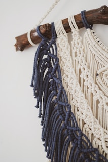 Detailed macram&eacute; wall hanging featuring dark blue and cream-colored cords intricately knotted. These are attached to a smooth wooden branch, creating a decorative piece with an artisanal feel.