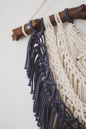 Detailed macramé wall hanging featuring dark blue and cream-colored cords intricately knotted. These are attached to a smooth wooden branch, creating a decorative piece with an artisanal feel.