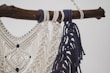 A detailed macramé design crafted with cream and navy blue threads, intricately knotted and hanging from a rustic wooden branch. The pattern features diamond shapes and decorative fringes.