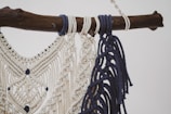 A detailed macramé design crafted with cream and navy blue threads, intricately knotted and hanging from a rustic wooden branch. The pattern features diamond shapes and decorative fringes.