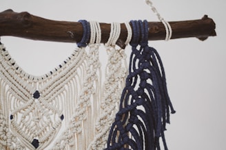 A detailed macramé design crafted with cream and navy blue threads, intricately knotted and hanging from a rustic wooden branch. The pattern features diamond shapes and decorative fringes.
