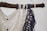 A detailed macramé design crafted with cream and navy blue threads, intricately knotted and hanging from a rustic wooden branch. The pattern features diamond shapes and decorative fringes.