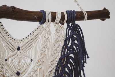 A detailed macramé design crafted with cream and navy blue threads, intricately knotted and hanging from a rustic wooden branch. The pattern features diamond shapes and decorative fringes.