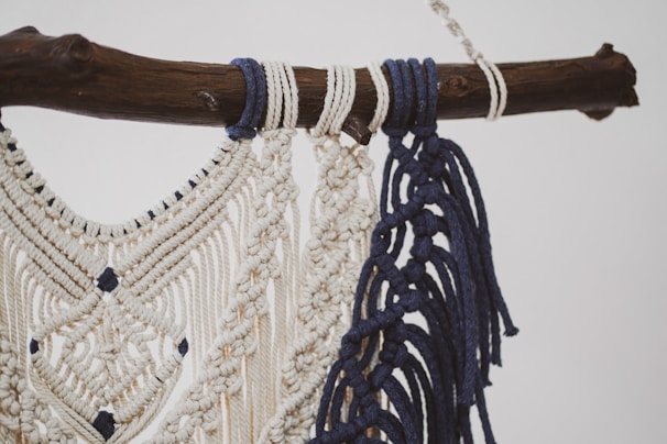 A detailed macramé design crafted with cream and navy blue threads, intricately knotted and hanging from a rustic wooden branch. The pattern features diamond shapes and decorative fringes.
