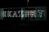 Large neon letters illuminated against a dark background, arranged to spell out a word in a modern, sans-serif font. Glass panels and curtains are visible behind the signage, suggesting an urban setting.