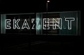 Large neon letters illuminated against a dark background, arranged to spell out a word in a modern, sans-serif font. Glass panels and curtains are visible behind the signage, suggesting an urban setting.