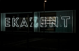 Large neon letters illuminated against a dark background, arranged to spell out a word in a modern, sans-serif font. Glass panels and curtains are visible behind the signage, suggesting an urban setting.