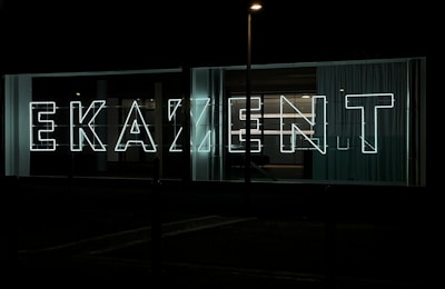Large neon letters illuminated against a dark background, arranged to spell out a word in a modern, sans-serif font. Glass panels and curtains are visible behind the signage, suggesting an urban setting.