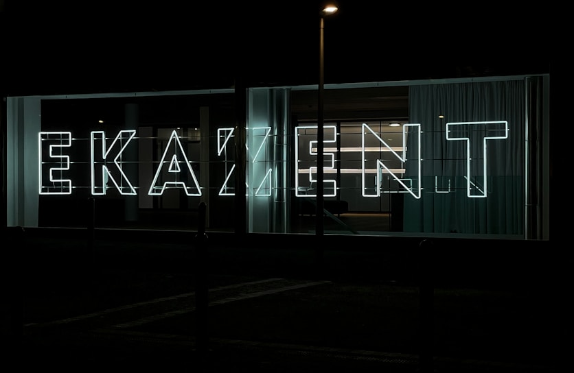 Large neon letters illuminated against a dark background, arranged to spell out a word in a modern, sans-serif font. Glass panels and curtains are visible behind the signage, suggesting an urban setting.