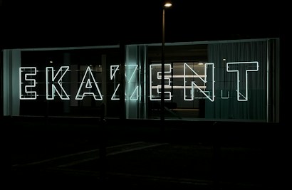 Large neon letters illuminated against a dark background, arranged to spell out a word in a modern, sans-serif font. Glass panels and curtains are visible behind the signage, suggesting an urban setting.
