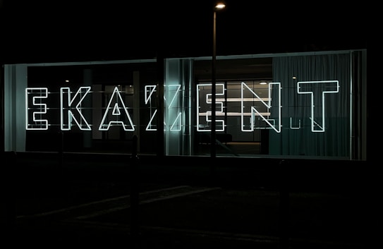 Large neon letters illuminated against a dark background, arranged to spell out a word in a modern, sans-serif font. Glass panels and curtains are visible behind the signage, suggesting an urban setting.