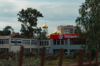 A shopping center with a prominent red and white sign featuring Cyrillic letters is in the foreground. Behind the building, golden onion domes of a church are visible. Trees and a multi-story apartment building are also in the background.