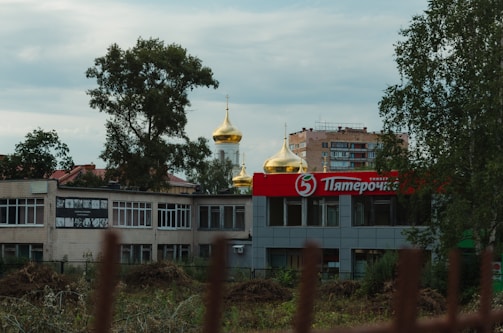 A shopping center with a prominent red and white sign featuring Cyrillic letters is in the foreground. Behind the building, golden onion domes of a church are visible. Trees and a multi-story apartment building are also in the background.