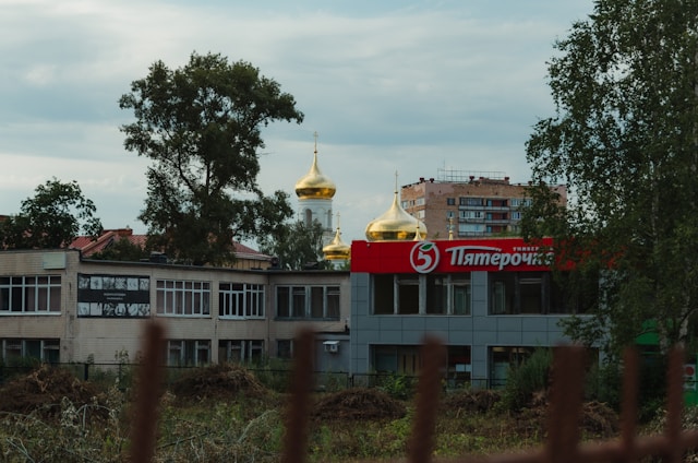 A shopping center with a prominent red and white sign featuring Cyrillic letters is in the foreground. Behind the building, golden onion domes of a church are visible. Trees and a multi-story apartment building are also in the background.