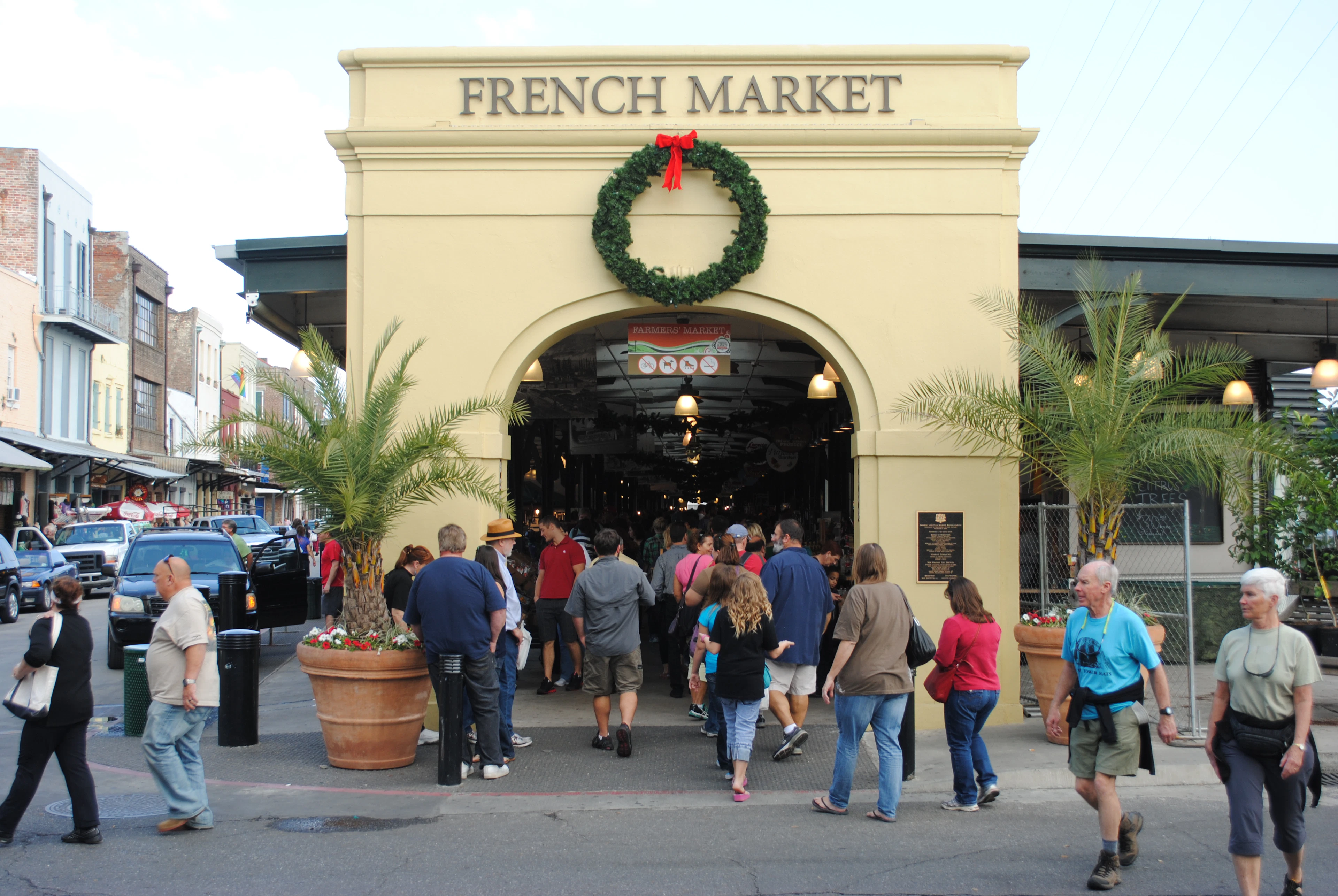 The pale yellow arched entrance of the French Market in New Orleans, its name carved above the doorway and a wreath hung beneath, with visitors streaming into the covered hall between potted palms in soft evening light.