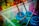 A collection of paint jars in various vibrant colors, including blue, green, and pink. Wooden paintbrushes covered in paint rest inside the jars, and the splattered paint around them suggests creative activity. The scene is set in a transparent plastic container.