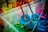 A collection of paint jars in various vibrant colors, including blue, green, and pink. Wooden paintbrushes covered in paint rest inside the jars, and the splattered paint around them suggests creative activity. The scene is set in a transparent plastic container.
