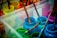 A collection of paint jars in various vibrant colors, including blue, green, and pink. Wooden paintbrushes covered in paint rest inside the jars, and the splattered paint around them suggests creative activity. The scene is set in a transparent plastic container.