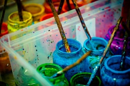 A collection of paint jars in various vibrant colors, including blue, green, and pink. Wooden paintbrushes covered in paint rest inside the jars, and the splattered paint around them suggests creative activity. The scene is set in a transparent plastic container.
