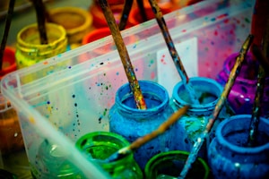 A collection of paint jars in various vibrant colors, including blue, green, and pink. Wooden paintbrushes covered in paint rest inside the jars, and the splattered paint around them suggests creative activity. The scene is set in a transparent plastic container.