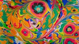 A vibrant and colorful abstract artwork featuring various swirling patterns and shapes. The colors blend together in a marbled effect, with floral motifs scattered throughout the image. The design includes bright shades of green, orange, yellow, pink, blue, black, and white.