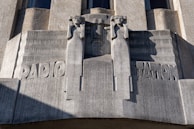 A sculptural relief on a building facade features two stylized human figures flanking the word 'RADIO STATION.' The figures have an angular, geometric design and appear to be holding their arms up. The material of the sculpture is a gray stone with visible texture and shadows cast by the sun.