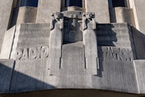 A sculptural relief on a building facade features two stylized human figures flanking the word 'RADIO STATION.' The figures have an angular, geometric design and appear to be holding their arms up. The material of the sculpture is a gray stone with visible texture and shadows cast by the sun.