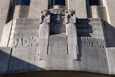 A sculptural relief on a building facade features two stylized human figures flanking the word 'RADIO STATION.' The figures have an angular, geometric design and appear to be holding their arms up. The material of the sculpture is a gray stone with visible texture and shadows cast by the sun.