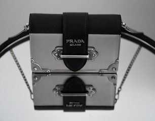 A stylish handbag with a metallic and leather design, featuring the Prada brand name on the front. The bag is elegantly placed on a reflective surface, creating a mirrored effect. The leather strap is prominently visible, with metal embellishments adding to the luxurious feel.