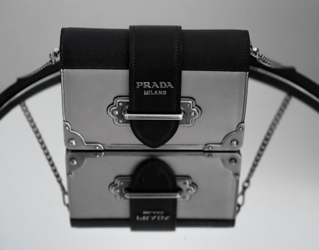 A stylish handbag with a metallic and leather design, featuring the Prada brand name on the front. The bag is elegantly placed on a reflective surface, creating a mirrored effect. The leather strap is prominently visible, with metal embellishments adding to the luxurious feel.
