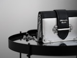 A luxury handbag with a sleek design featuring a metallic silver and black color scheme, adorned with a prominent logo. The bag is placed on a reflective surface, enhancing its elegant and modern appeal.