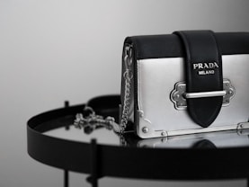 A luxury handbag with a sleek design featuring a metallic silver and black color scheme, adorned with a prominent logo. The bag is placed on a reflective surface, enhancing its elegant and modern appeal.