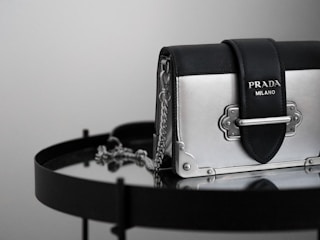 A luxury handbag with a sleek design featuring a metallic silver and black color scheme, adorned with a prominent logo. The bag is placed on a reflective surface, enhancing its elegant and modern appeal.