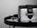 A luxury handbag with a sleek design featuring a metallic silver and black color scheme, adorned with a prominent logo. The bag is placed on a reflective surface, enhancing its elegant and modern appeal.