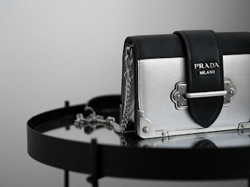 A luxury handbag with a sleek design featuring a metallic silver and black color scheme, adorned with a prominent logo. The bag is placed on a reflective surface, enhancing its elegant and modern appeal.