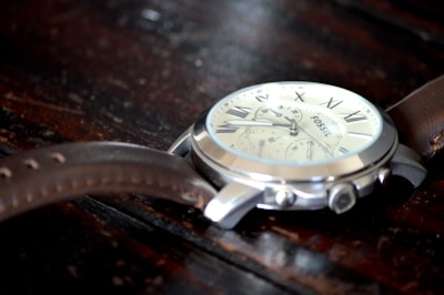 A close-up view of a wristwatch with a metallic casing and a brown leather strap. The watch face features Roman numerals and several small dials indicating additional timekeeping functions. The background is a textured dark wooden surface, adding contrast to the shiny and sleek appearance of the watch.