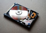 An open hard drive with its internal components visible, resting on a flat surface. The shiny, reflective metal platter is prominent, surrounded by the actuator arm and the read/write head, along with various colorful circuitry and connectors.