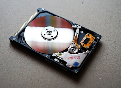 An open hard drive with its internal components visible, resting on a flat surface. The shiny, reflective metal platter is prominent, surrounded by the actuator arm and the read/write head, along with various colorful circuitry and connectors.