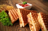 Golden brown waffle sandwiches are arranged on a wooden table, filled with a savory mixture that appears to include vegetables and cheese. A fresh lettuce leaf sits to one side, adding a touch of green. In the background, crispy potato wedges and a small dish of salsa or chutney provide additional colorful elements.
