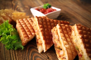 Golden brown waffle sandwiches are arranged on a wooden table, filled with a savory mixture that appears to include vegetables and cheese. A fresh lettuce leaf sits to one side, adding a touch of green. In the background, crispy potato wedges and a small dish of salsa or chutney provide additional colorful elements.