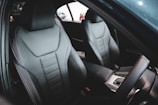 The interior of a car focusing on two front seats. Both seats are upholstered in black leather and feature detailed stitching and ergonomic contours. The image also shows part of the steering wheel and a glimpse of the center console.