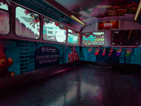 The interior of a bus or vehicle is decorated with vibrant holiday themes, featuring a 'Genting Winter Wonderland' banner. The walls display illustrations of teddy bears, colorful Christmas stockings, and gifts. The windows are adorned with festive garlands, creating a cheerful holiday atmosphere.