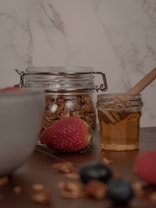 A rustic table scene featuring a glass jar filled with granola or cereal, a small clear jar containing honey with a wooden honey dipper, and assorted berries like strawberries and blueberries scattered around. The background is a light marble texture, adding a clean and natural feel to the composition.