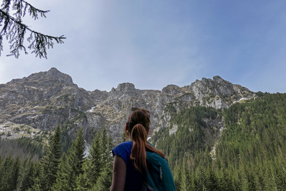 woman in blue shirt standing near green trees and mountain during daytime