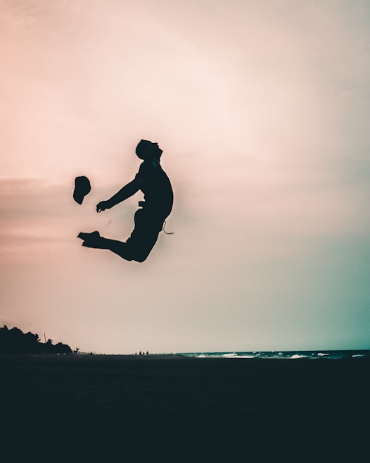 A silhouetted person is captured mid-air, performing a jump against a beach backdrop under a cloudy sky. The sea is visible in the distance, and a hat is airborne beside the person.