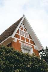 A charming house with a steep gable roof covered in dark terracotta tiles. The facade is adorned with geometric white trimmings against an orange-brown background. Lush greenery partially covers the lower part of the building.