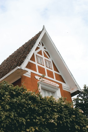 A charming house with a steep gable roof covered in dark terracotta tiles. The facade is adorned with geometric white trimmings against an orange-brown background. Lush greenery partially covers the lower part of the building.